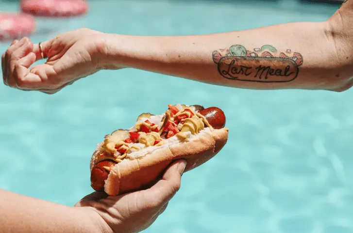 A Chicago style hot dog next to a person's arm that has a tattoo of a Chicago style hot dog. A swimming pool is the background.
