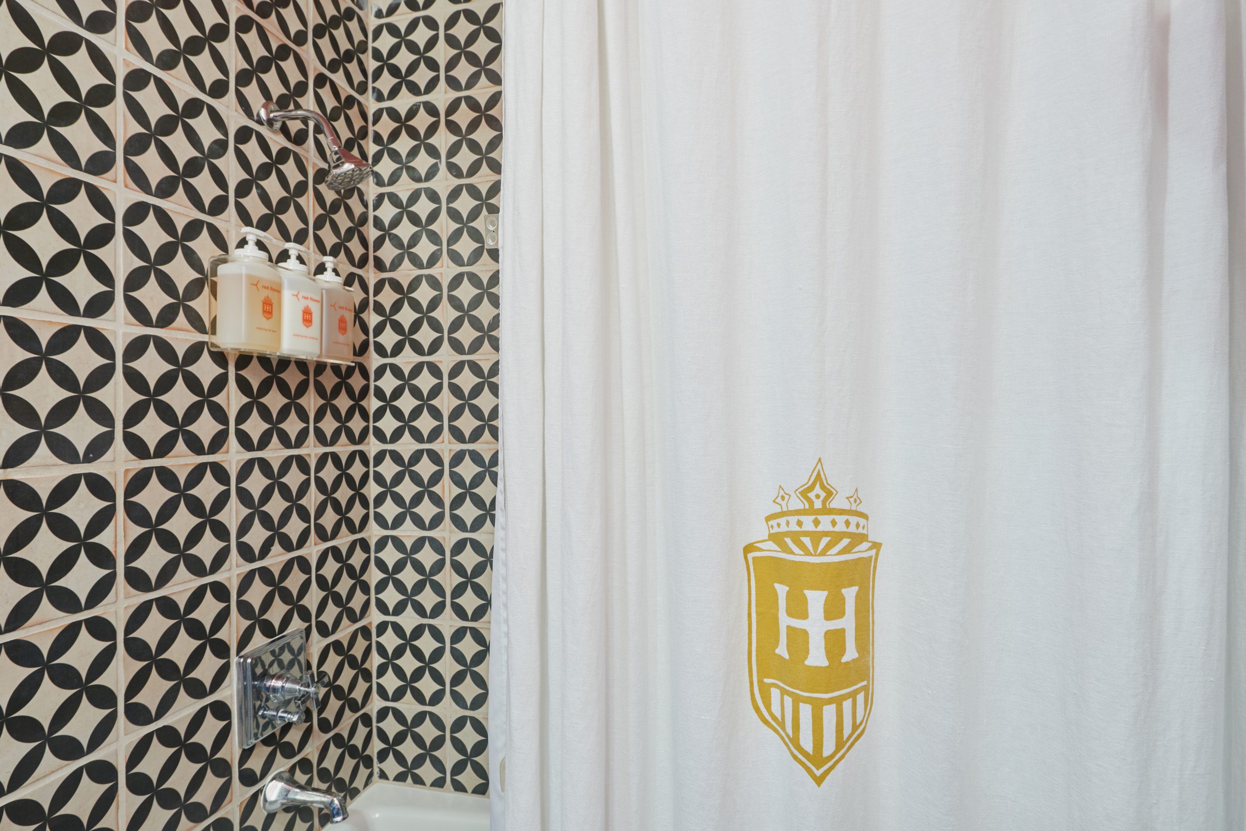 Black and white tiled shower with white shower curtain featuring gold Hotel Havana logo