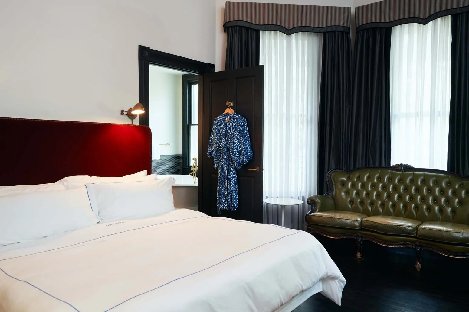 A green leather chair, a king sized bed, and black curtains with a blue robe hanging on a door inside Suite Three