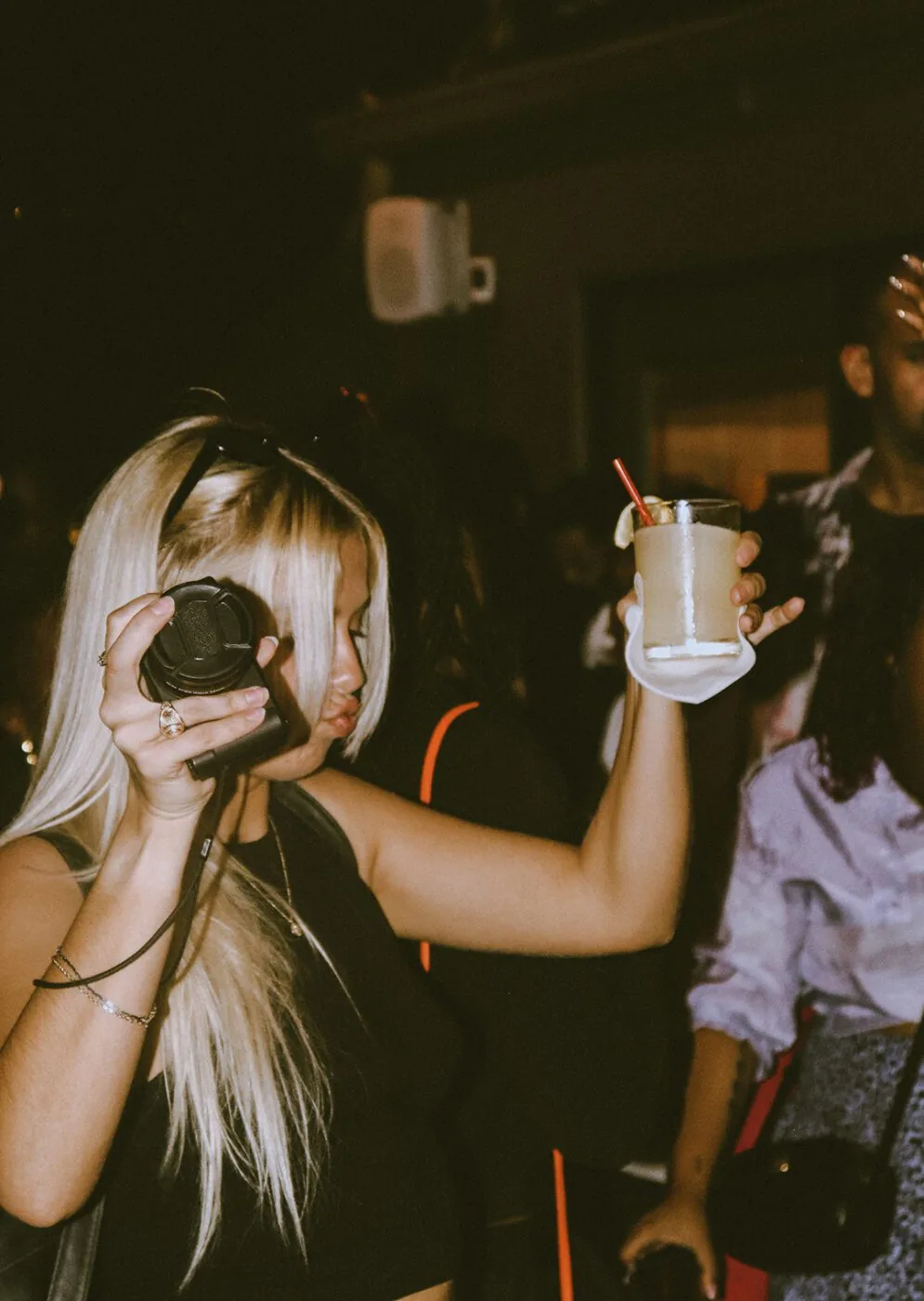 A blonde woman holding up a camera and a drink