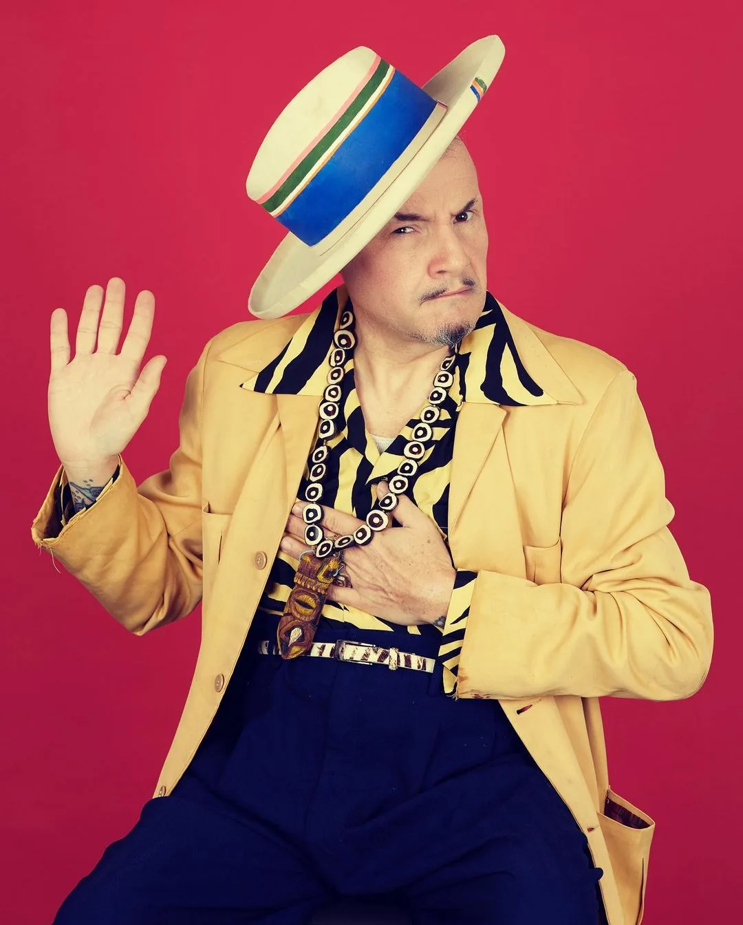 Man posing with one hand raised sitting against a pink background