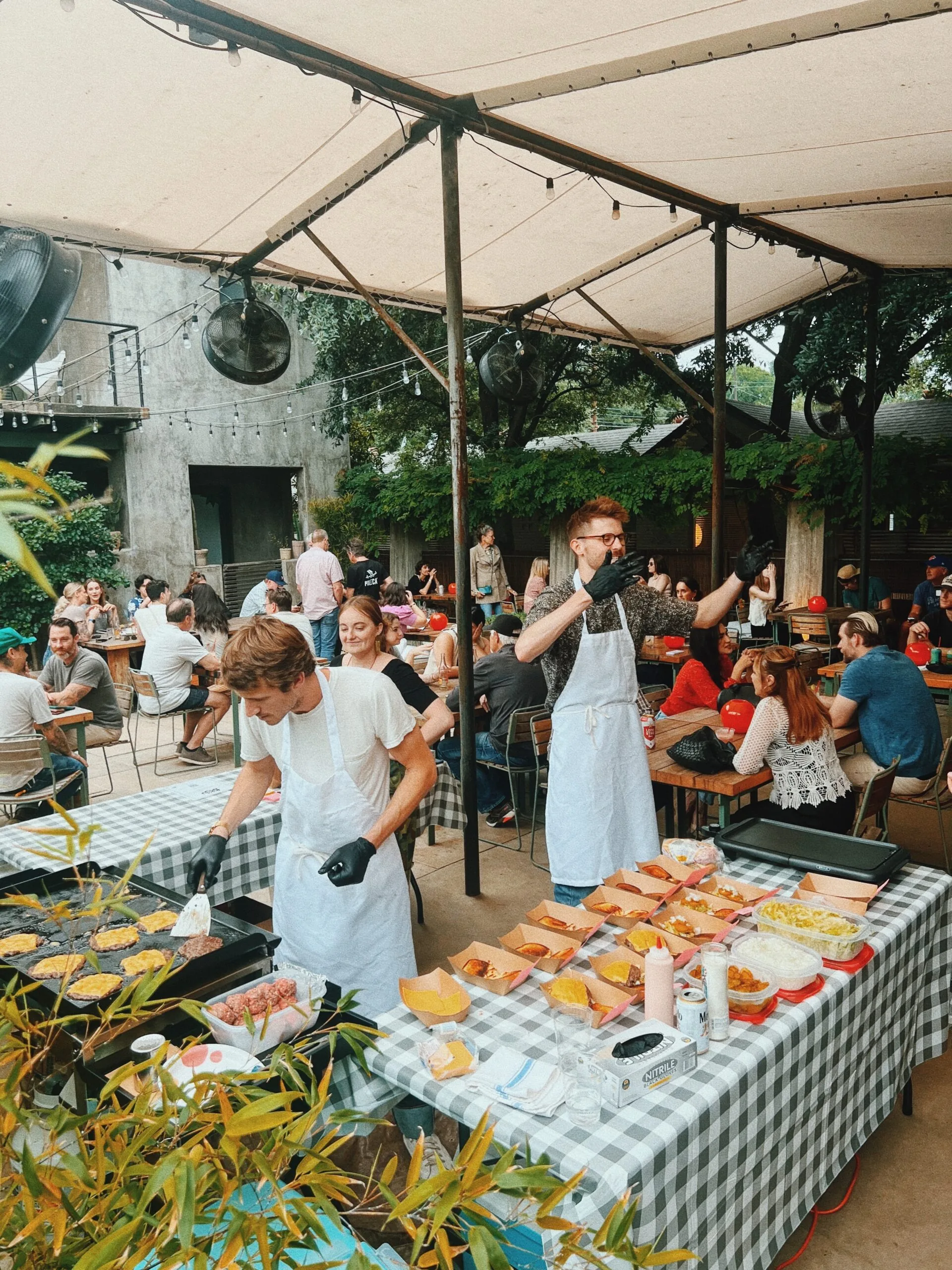 two men bbqing and preping food. surrounded by people seated at tables eating and talking