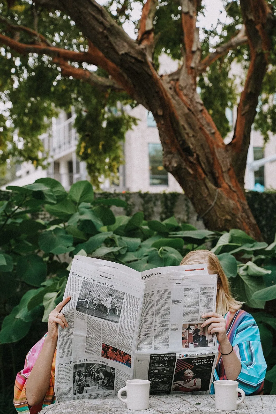 two people holding a newspaper up to read, sitting at a table with two mugs