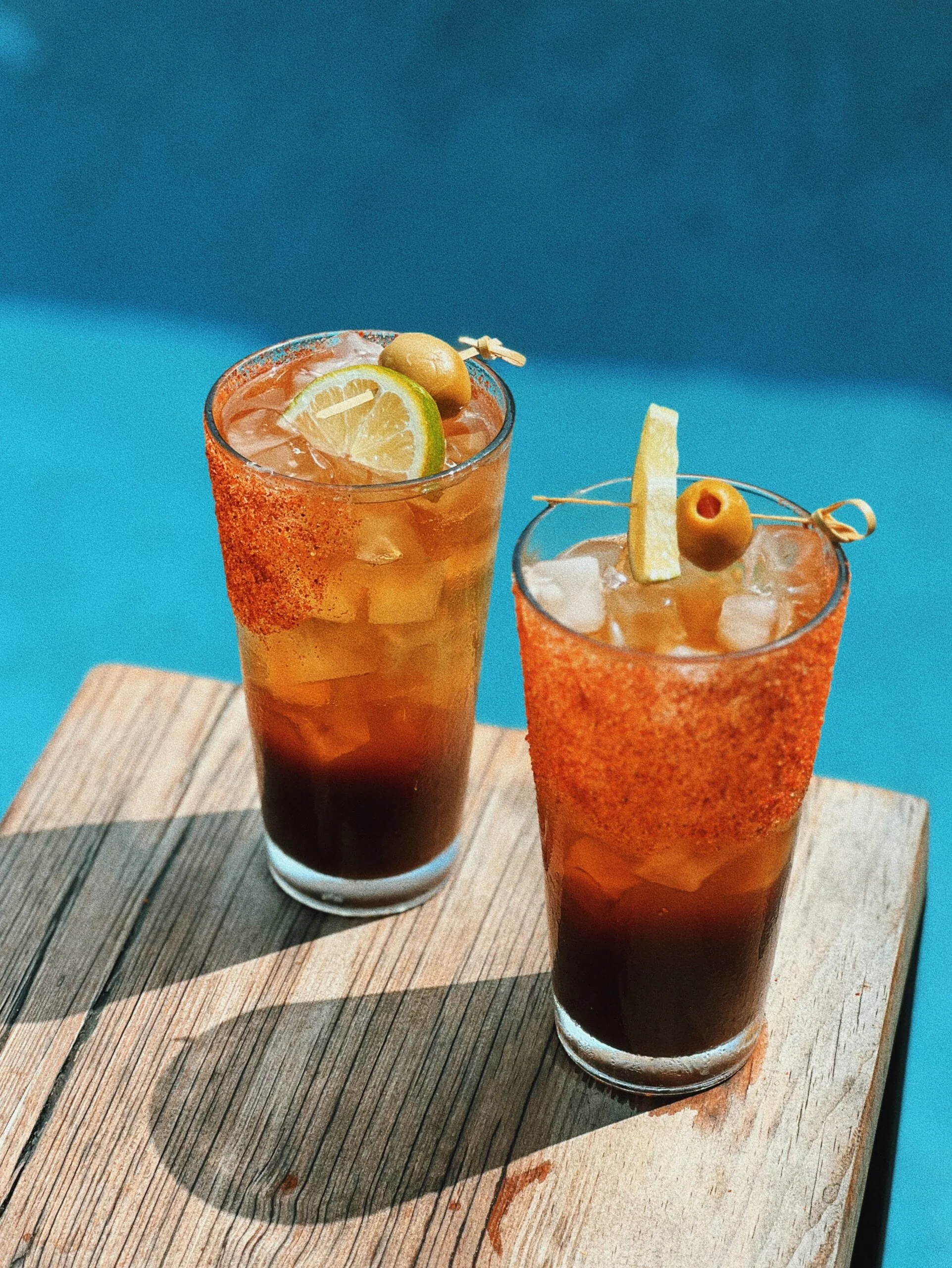 two glass cups filled with a drink topped with ice, olive and lemon. on a wooden table