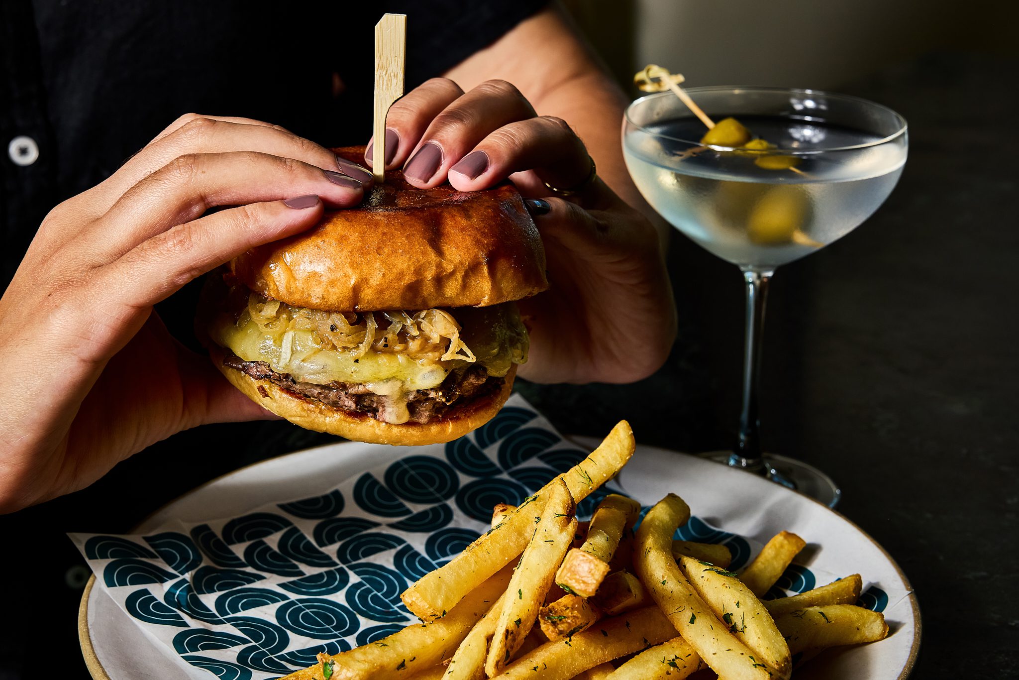 Handing holding a cheeseburger with French fries and a martini.