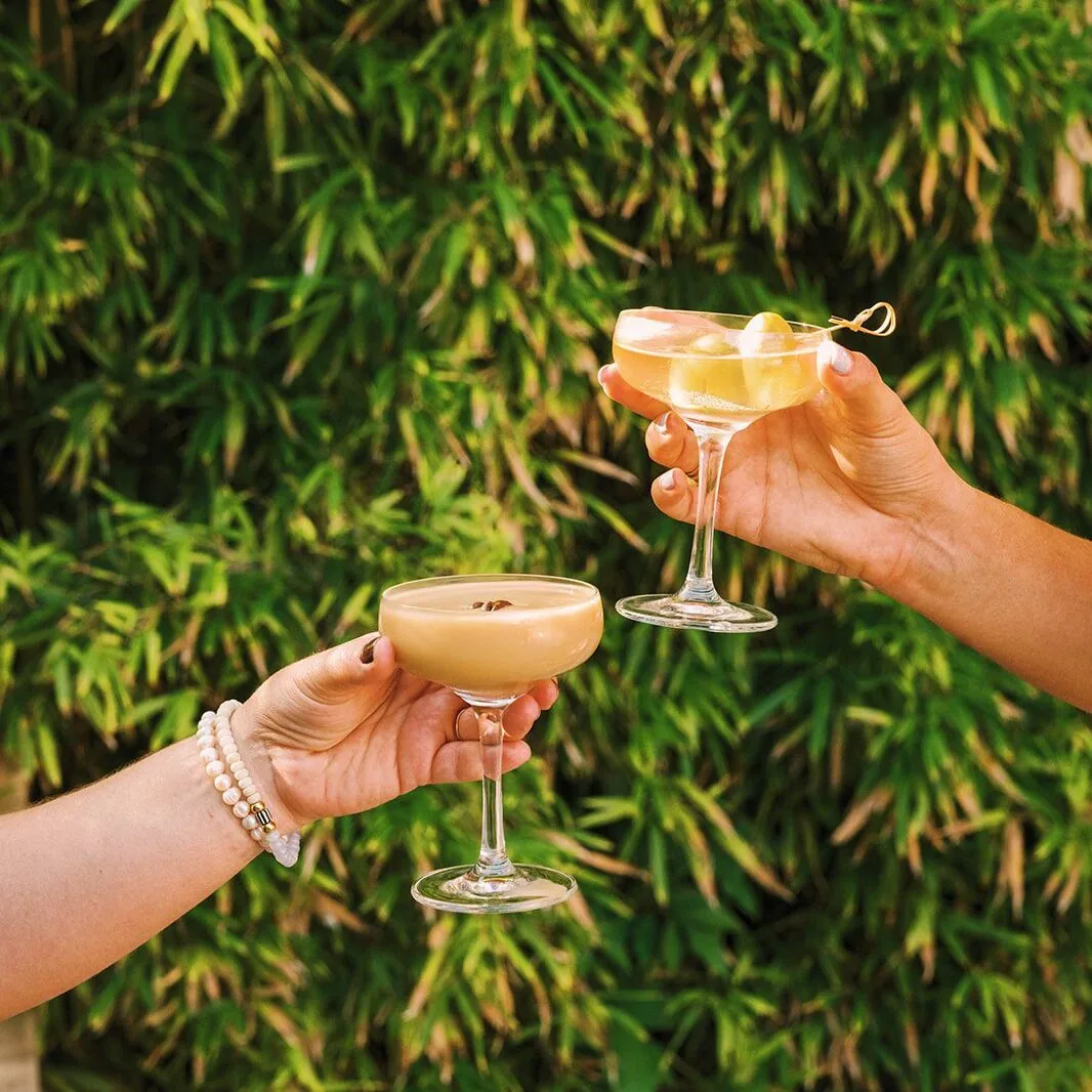 Two cocktails held in front of a wall of bamboo leaves